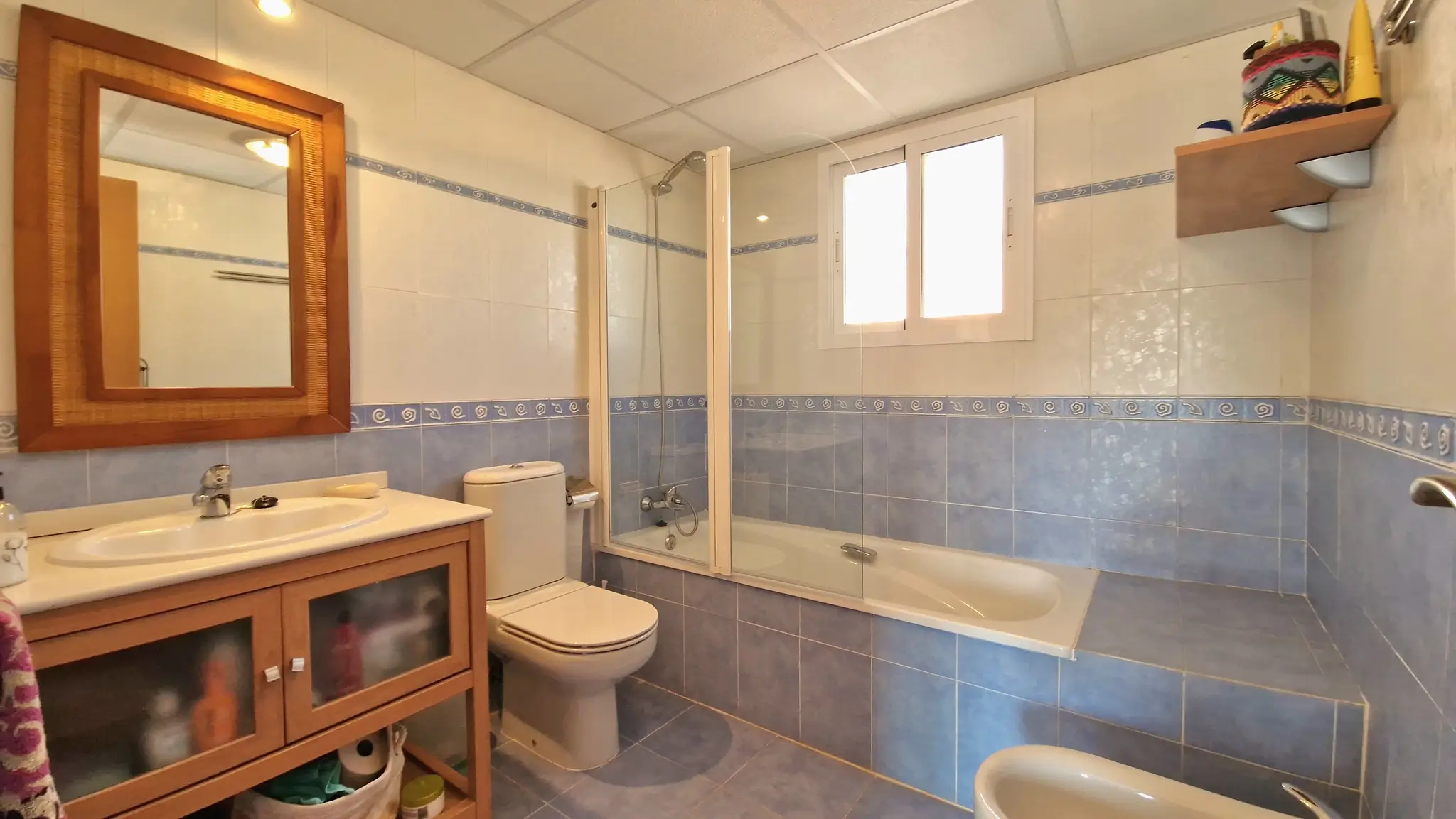 Bathroom featuring blue and white tiling, a wooden vanity with integrated sink, and a bathtub equipped with a glass screen and natural light from a window.