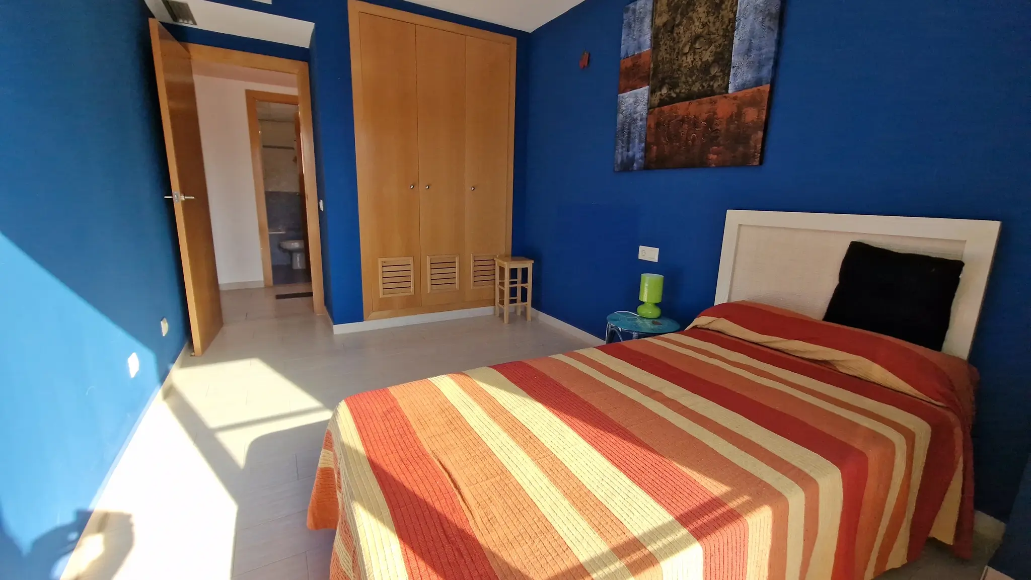 Bedroom with deep blue walls, built-in wooden wardrobe, and tiled floors. The space includes a single bed and provides direct access to an ensuite bathroom.
