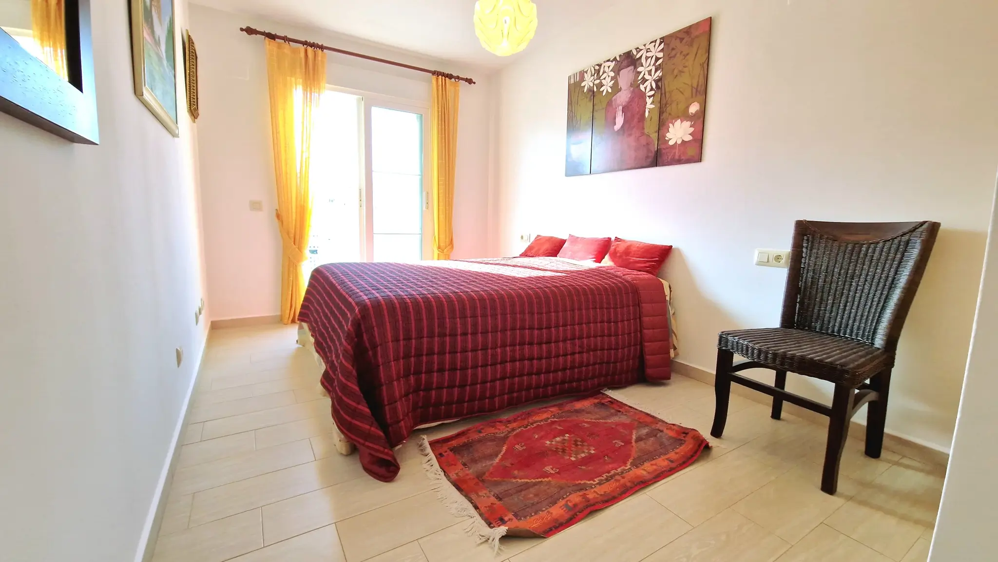 Bright bedroom featuring a double bed with red quilted bedding, yellow curtains, a dark wicker chair, and a decorative Buddha triptych on the wall.