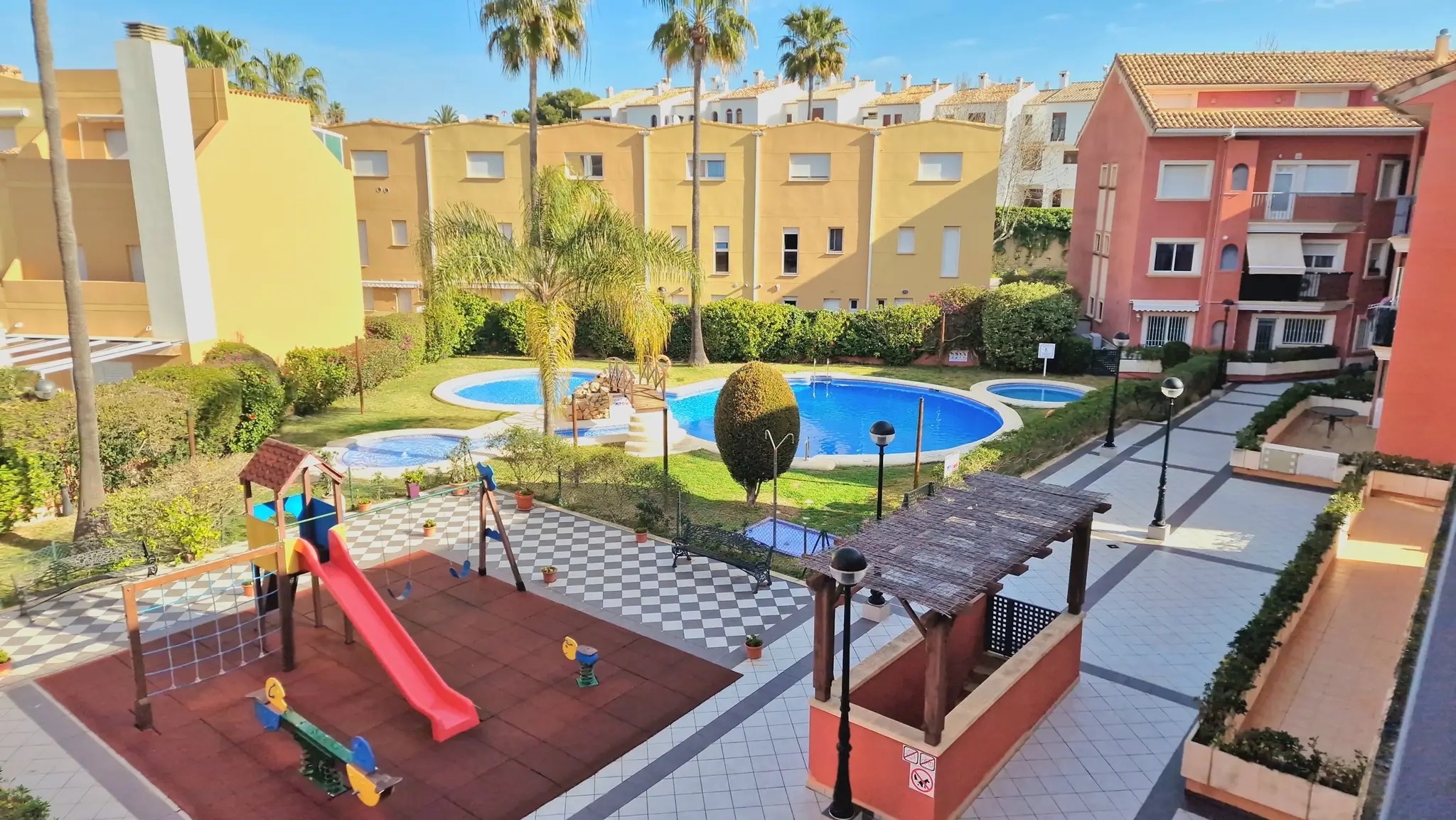 Communal area featuring a children's playground with safety flooring, a large swimming pool with a decorative bridge, and landscaped gardens set against Mediterranean-style buildings.