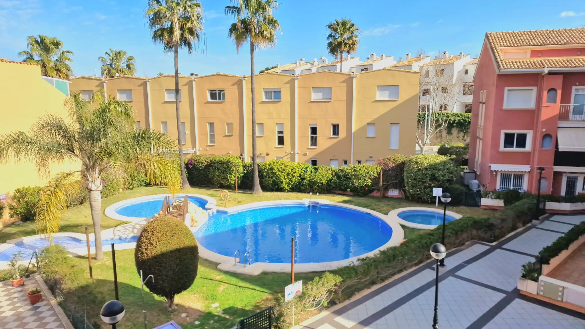 Communal area featuring a curved swimming pool, integrated children's pool, manicured lawns, and palm trees within a residential complex.