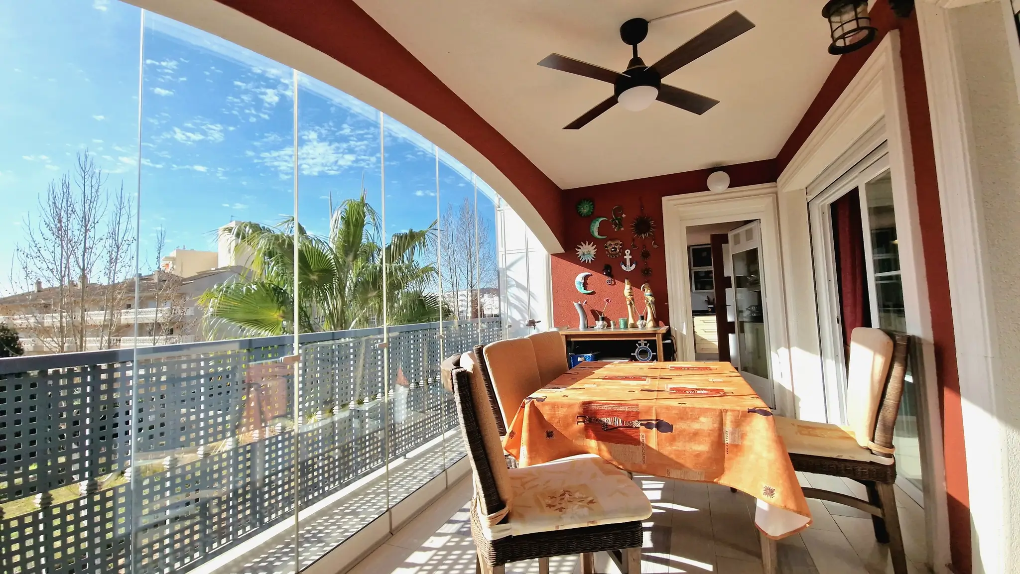 Covered terrace featuring frameless glass curtains, an outdoor dining area with wicker chairs, a ceiling fan, and views of palm trees and the sky.