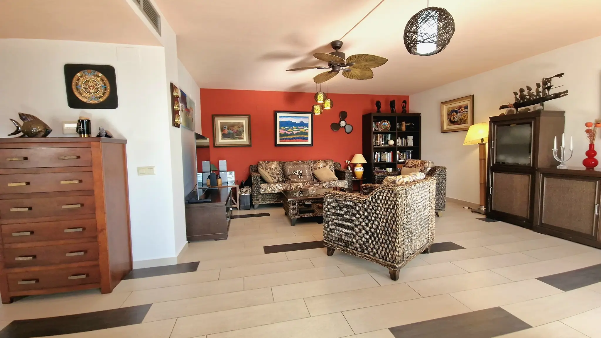Eclectic living room featuring wicker seating, a terracotta-red accent wall, a leaf-blade ceiling fan, and tiled flooring with dark decorative inserts.