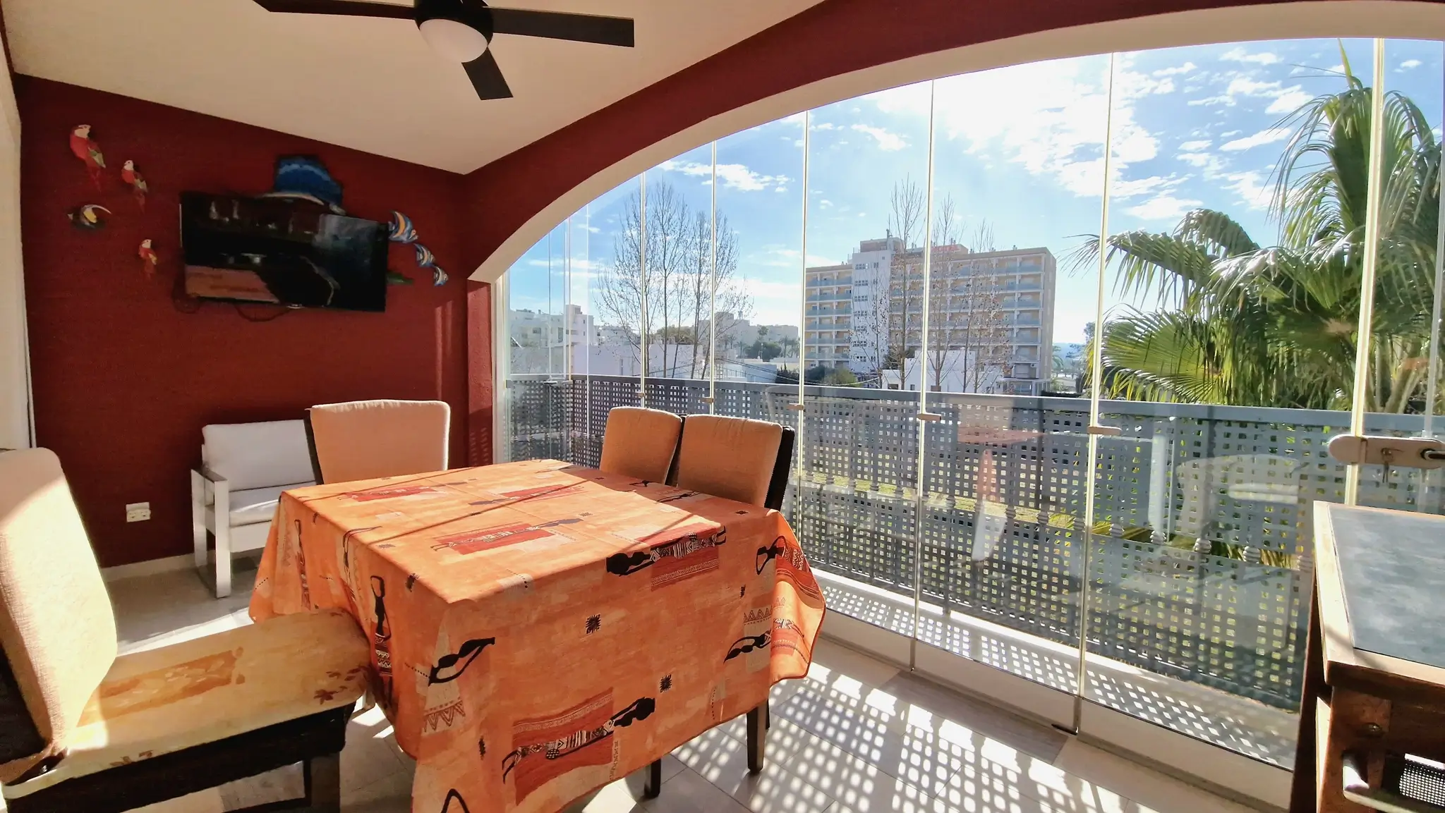 Enclosed terrace dining area featuring frameless glass curtains, a red accent wall with a wall-mounted TV, and urban views under bright natural light.