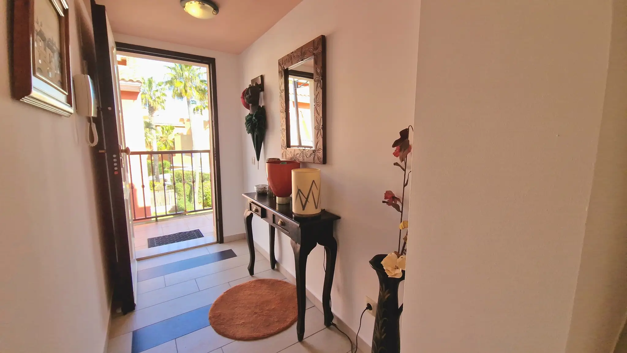Entryway featuring a dark wood console table, decorative mirror, and tiled flooring, with a balcony door offering views of palm trees and natural light.