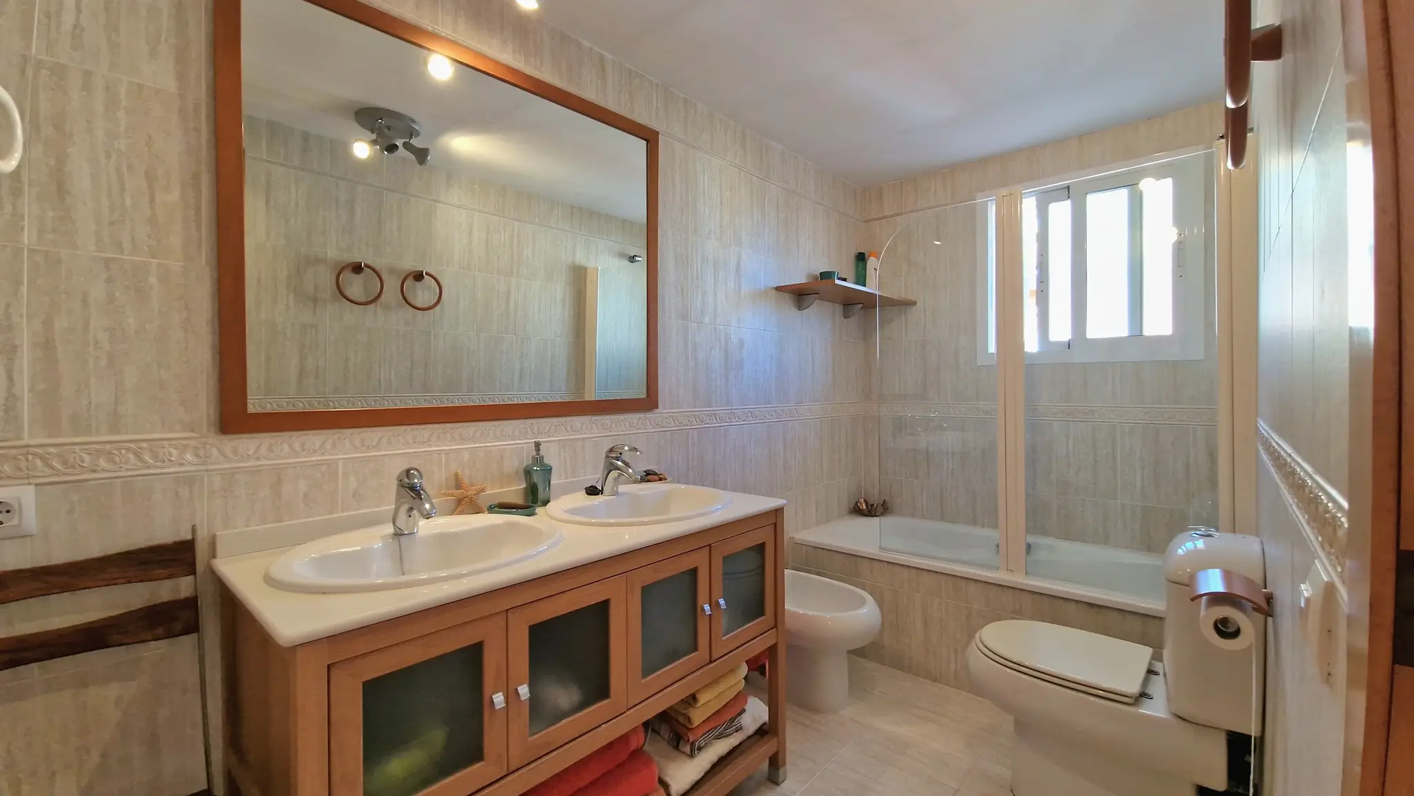 Full bathroom featuring a double vanity with wooden cabinetry, a large framed mirror, and a bidet. The space includes a bathtub with a glass screen and neutral tiled walls.