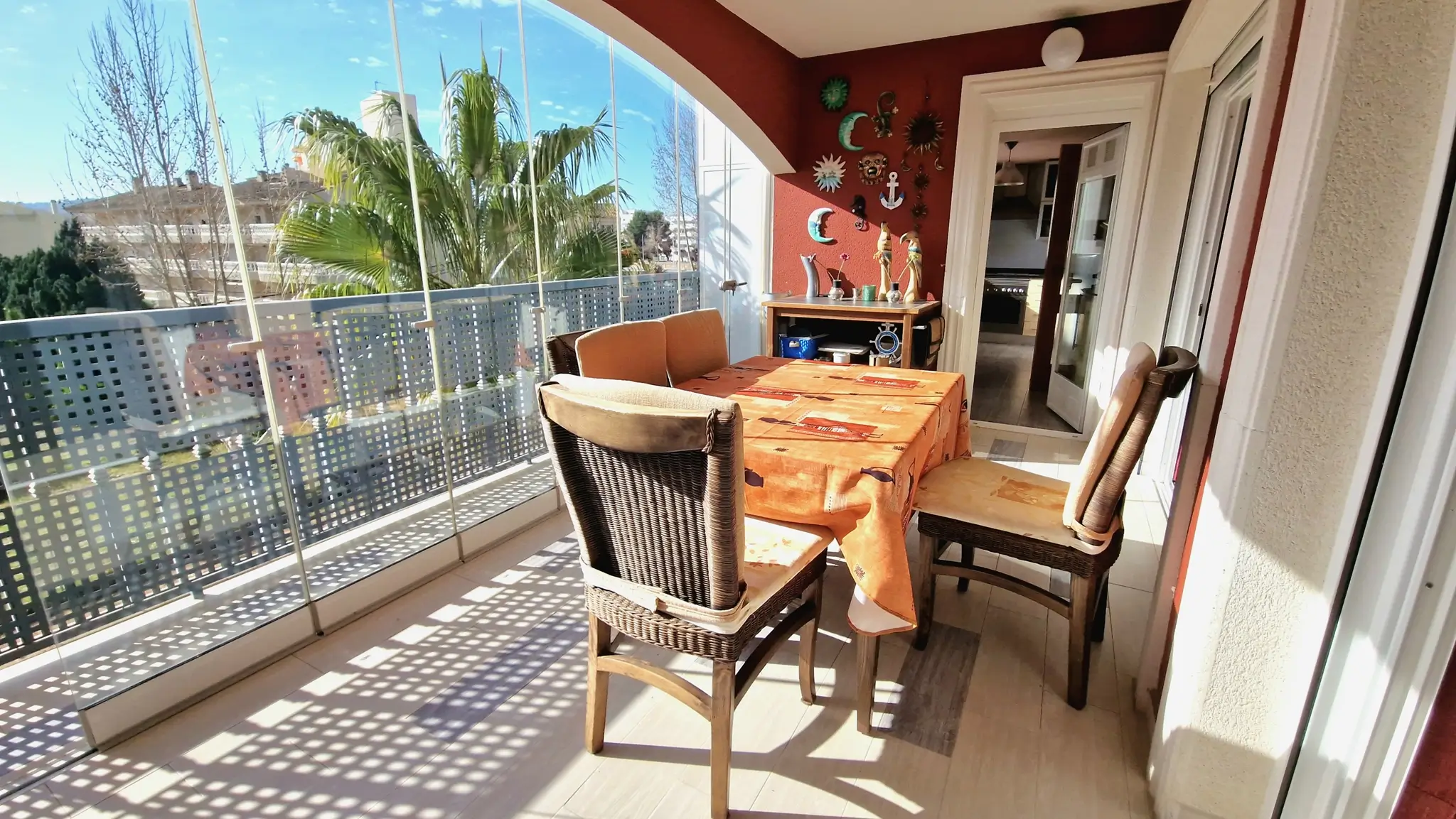 Glazed terrace featuring glass curtains, a dining area with wicker chairs, and a red accent wall with decorative art overlooking palm trees.