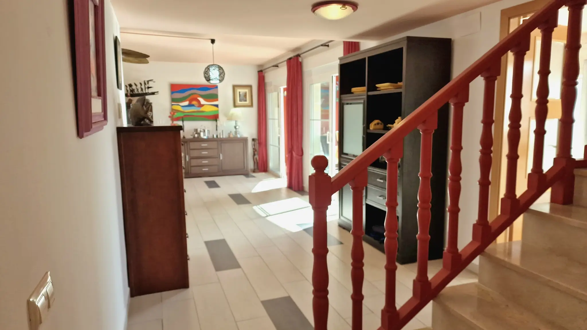 Hallway featuring a red wooden banister and marble stairs, leading to a bright living area with tiled floors, dark wood cabinetry, and vibrant red curtains.
