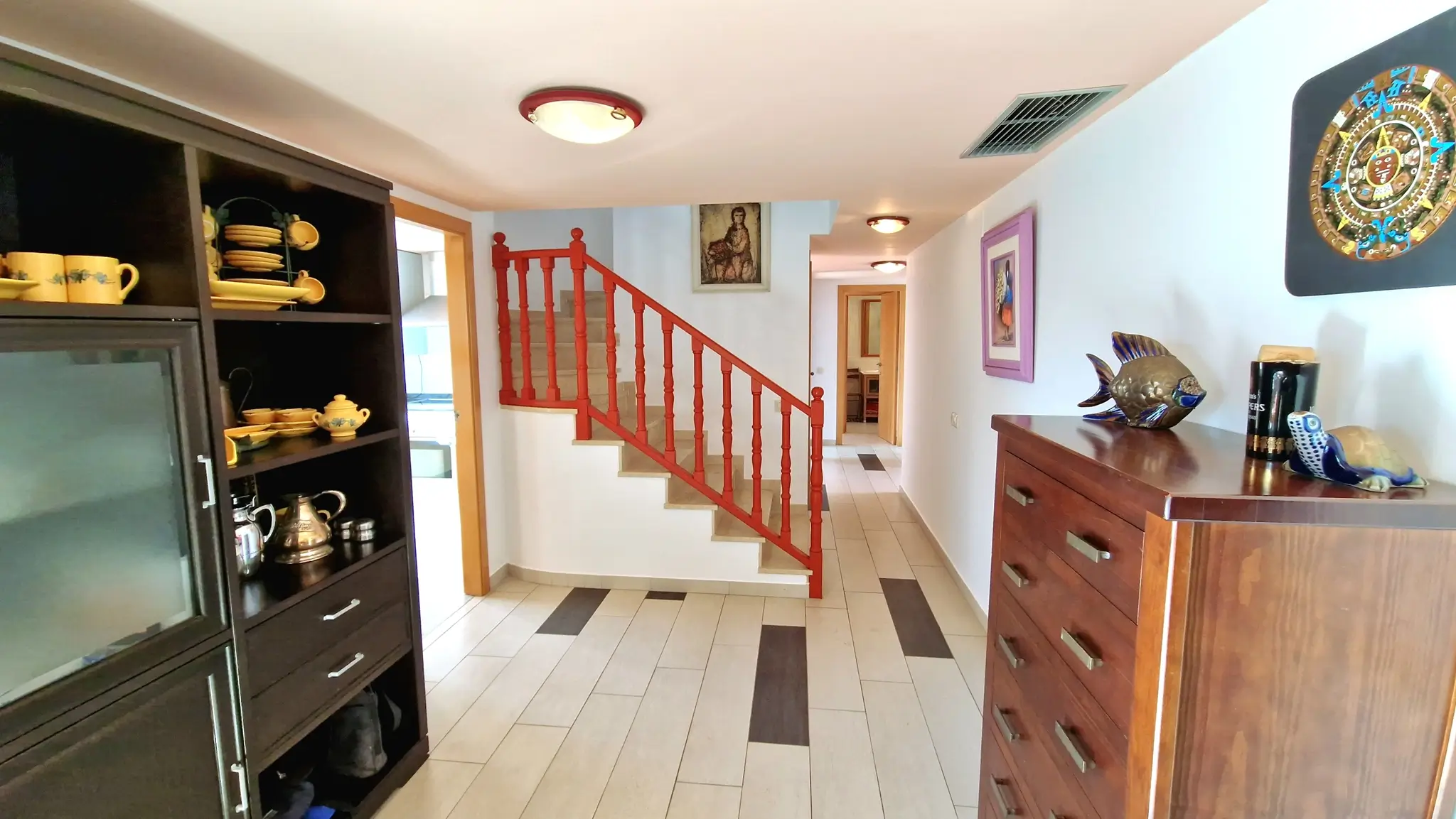 Hallway featuring a staircase with a red wooden railing, tiled flooring with dark accents, a dark wood display cabinet, and a wooden chest of drawers.