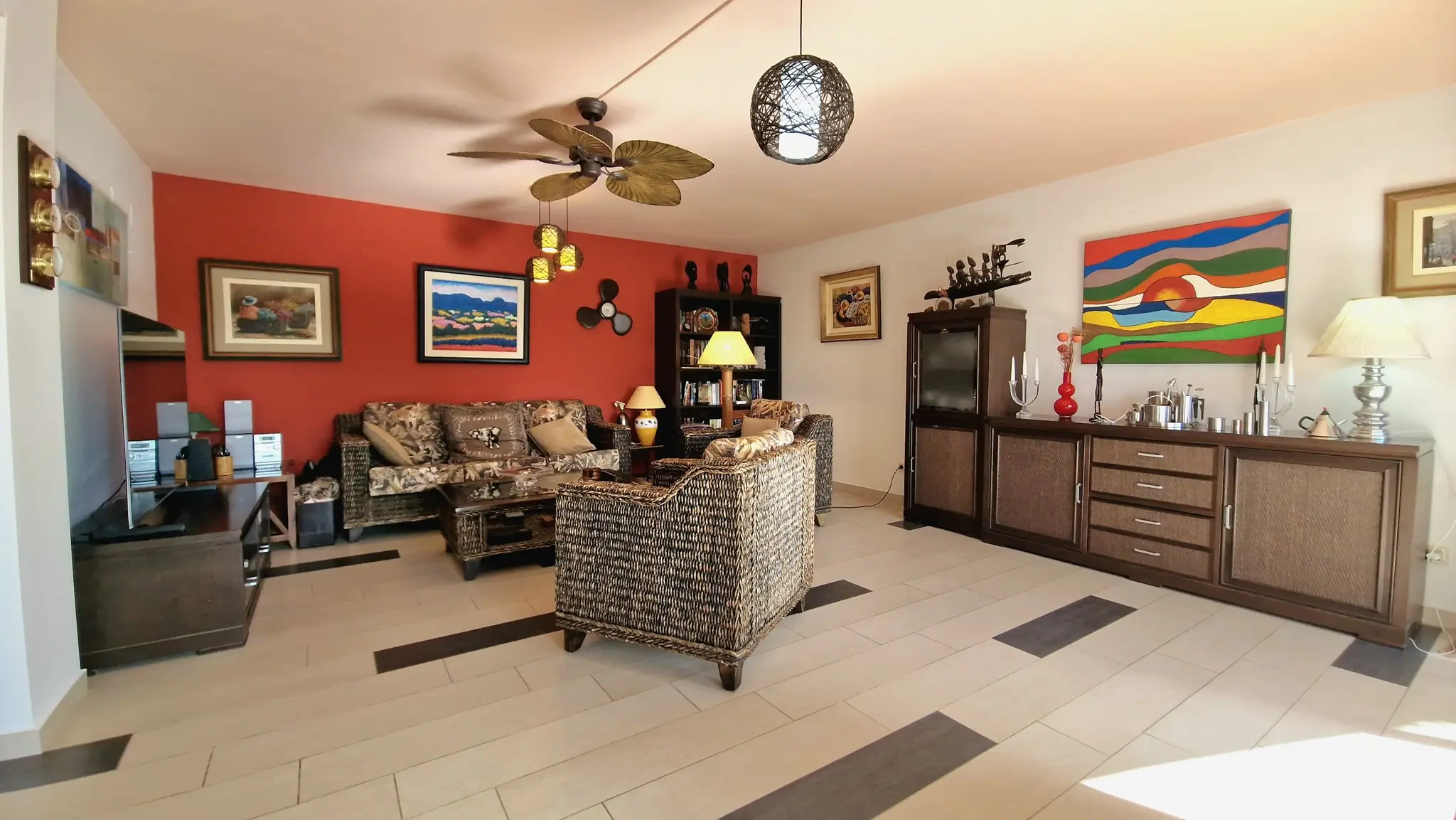 Living area featuring a red accent wall, wicker furniture, and a palm-leaf ceiling fan. The space includes tiled flooring with dark inlays and diverse art pieces.