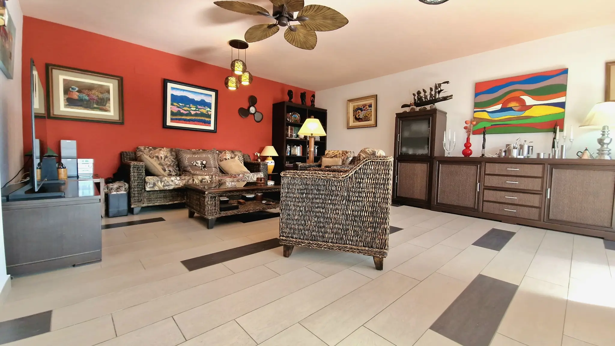 Living area featuring wicker furniture, a terracotta accent wall with framed art, and a leaf-style ceiling fan over tiled flooring with dark inserts.