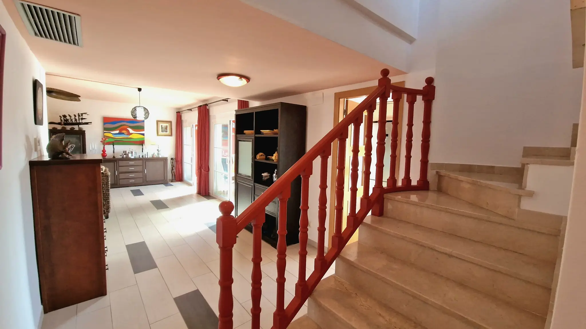 Marble staircase with a distinctive red wooden railing leading to a bright living area featuring tiled flooring and dark wood cabinetry.