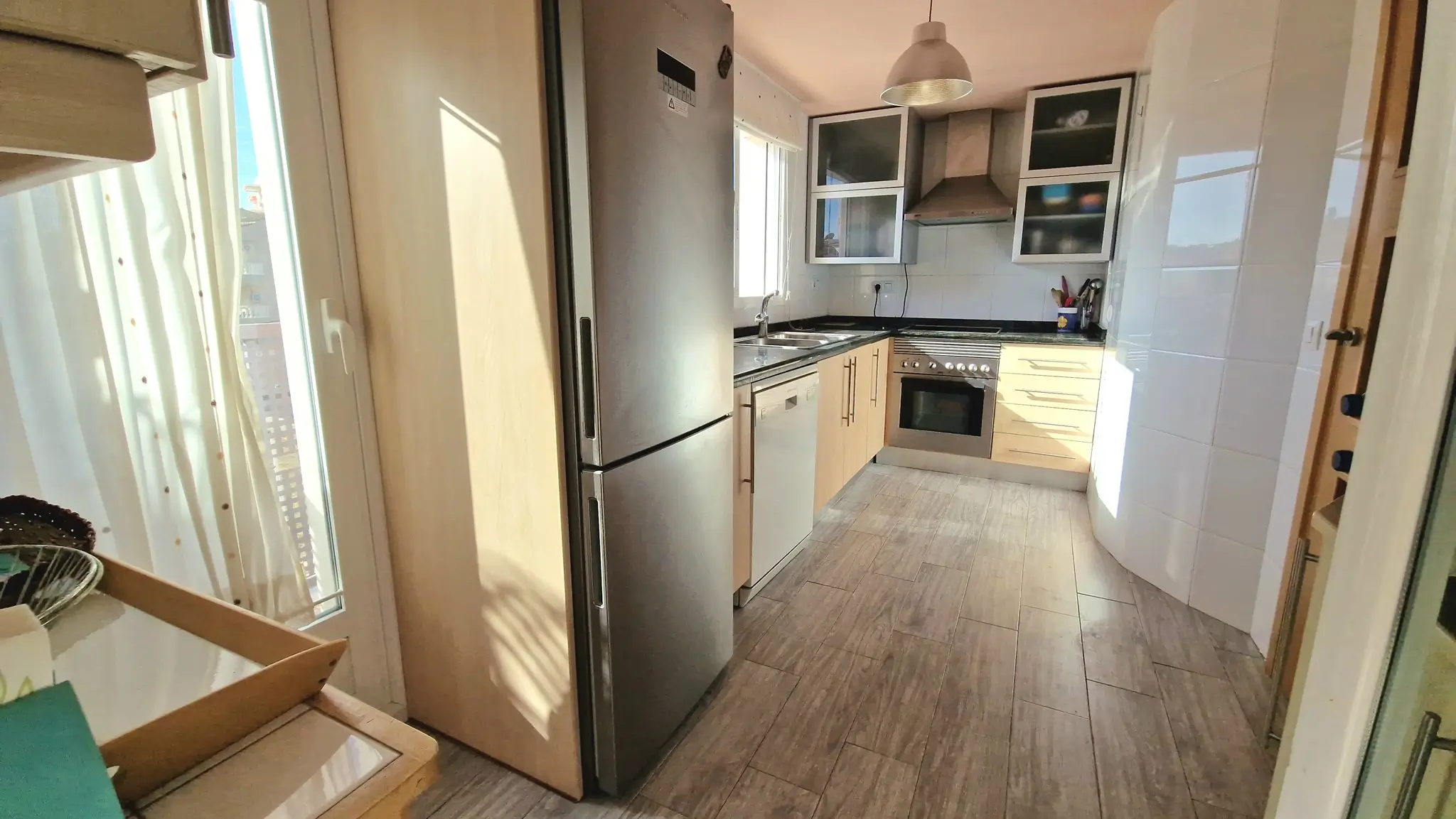 Modern galley kitchen featuring stainless steel appliances, light wood cabinetry, and wood-effect tile flooring illuminated by natural light.