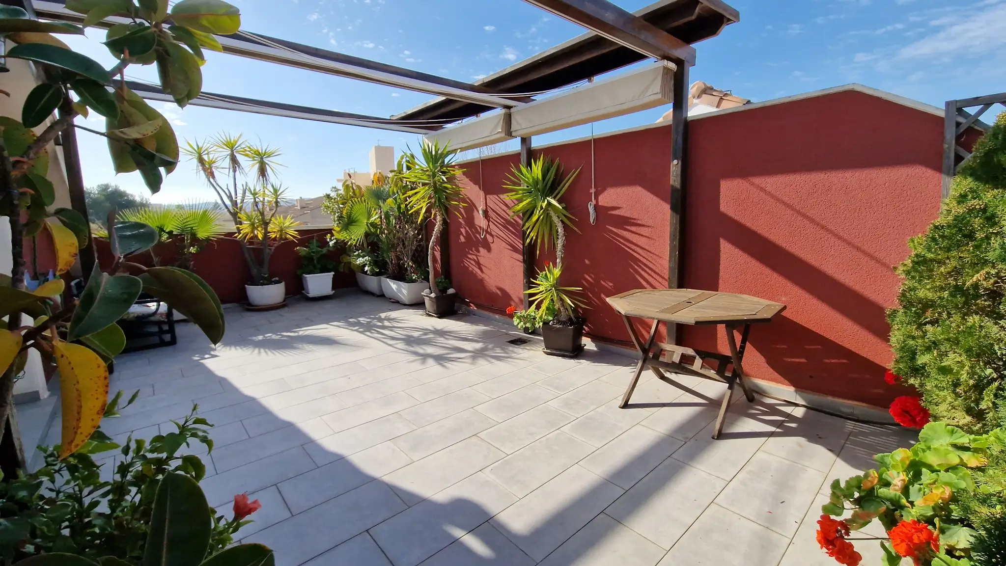 Outdoor terrace featuring grey floor tiles, a wooden pergola with a retractable awning, and various potted Mediterranean plants against a red wall.
