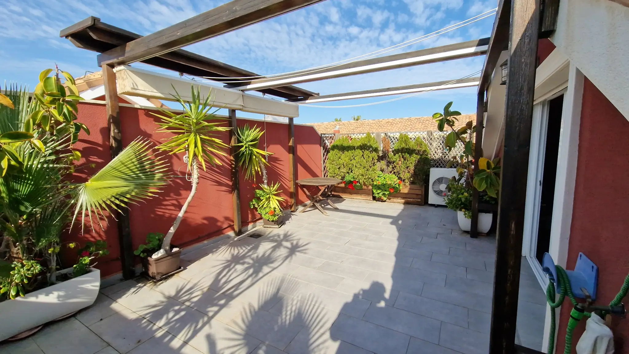 Private terrace with grey tiled flooring, a dark wood pergola, and retractable awnings. Features terracotta walls, various potted palms, and a small wooden table.