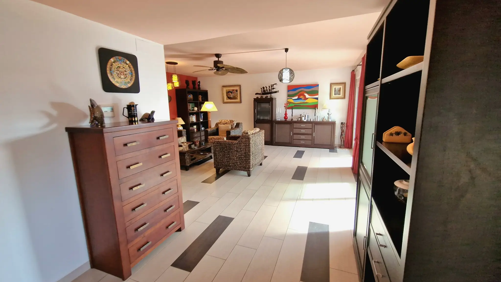 Spacious living area featuring light tiled flooring with dark accents, wicker armchairs, and a wooden chest of drawers under a ceiling fan.