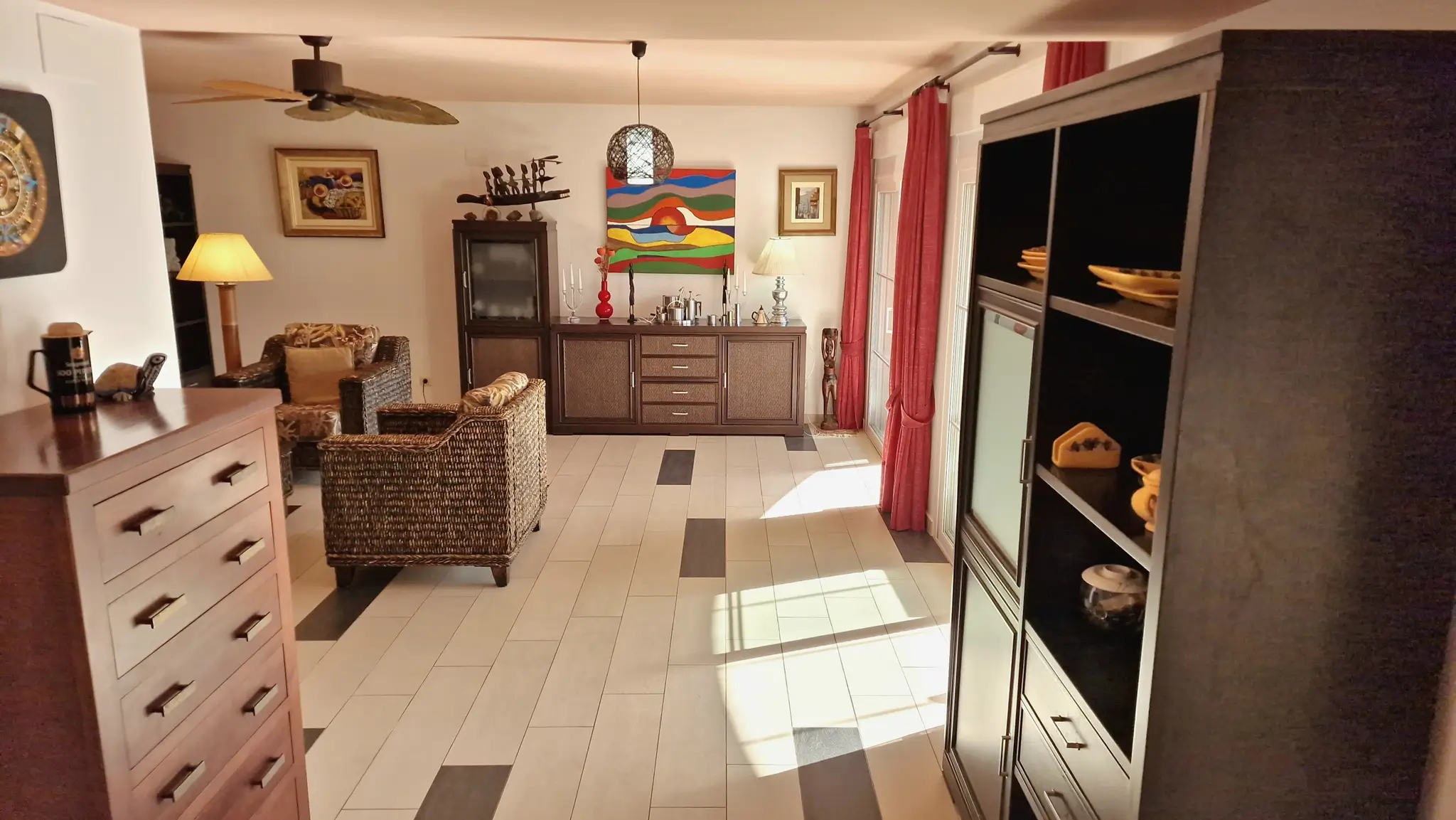Spacious living area featuring wicker armchairs, dark wood cabinetry, and tiled flooring with dark accents. Natural light enters through glass doors framed by red curtains.
