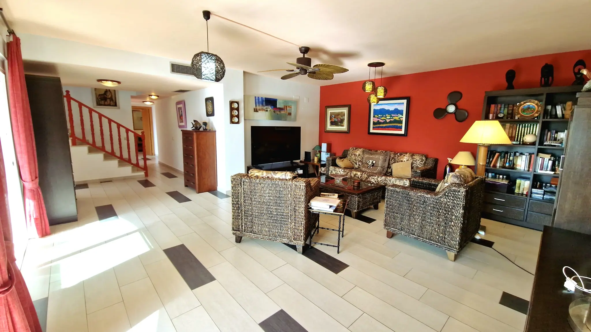 Spacious living room featuring a red accent wall, wicker furniture, and tiled flooring. Includes a staircase with red railings and a decorative leaf-blade ceiling fan.