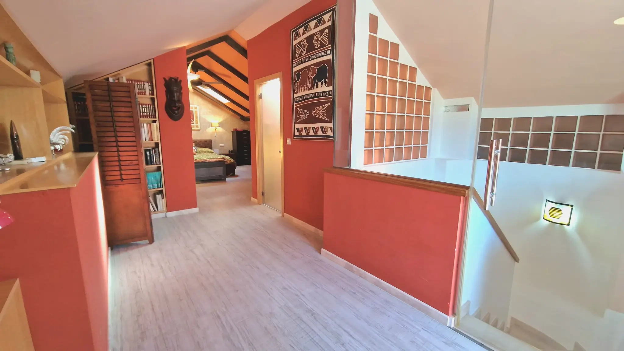 Upper-level landing with terracotta walls, light wood flooring, and glass railing. It leads to a bedroom featuring sloped ceilings with exposed dark wood beams and built-in shelving.