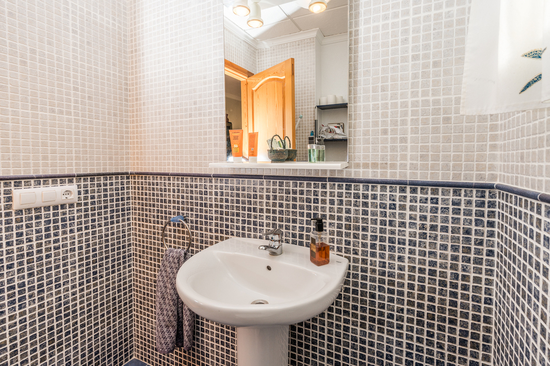 Bathroom featuring a white pedestal sink, mosaic tile walls in white and blue tones, and a mirror with integrated lighting and a small shelf.