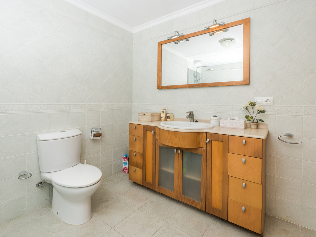 Bathroom featuring a wooden vanity with integrated sink, large wood-framed mirror with overhead lighting, and light-toned wall tiles with decorative borders.