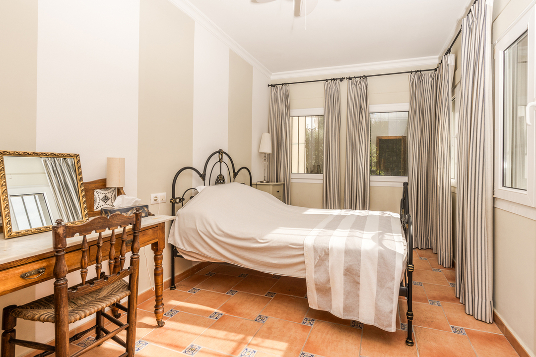 Bedroom featuring a black wrought iron bed, striped wallpaper, and terracotta flooring with decorative tile inserts. A classic wooden vanity desk sits beside the bed.