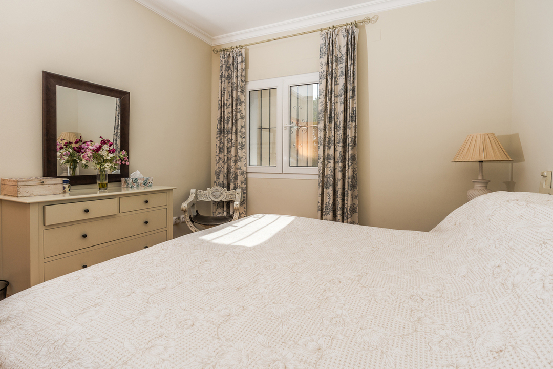 Bedroom featuring a double bed, a wooden dresser with a mirror, floral patterned curtains, and a window with security bars.