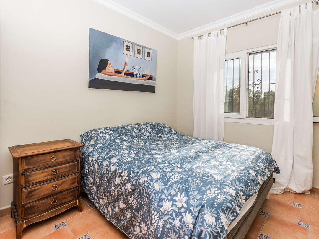 Bedroom featuring a double bed with blue floral bedding, a classic wooden nightstand, and terracotta floor tiles with decorative inserts.