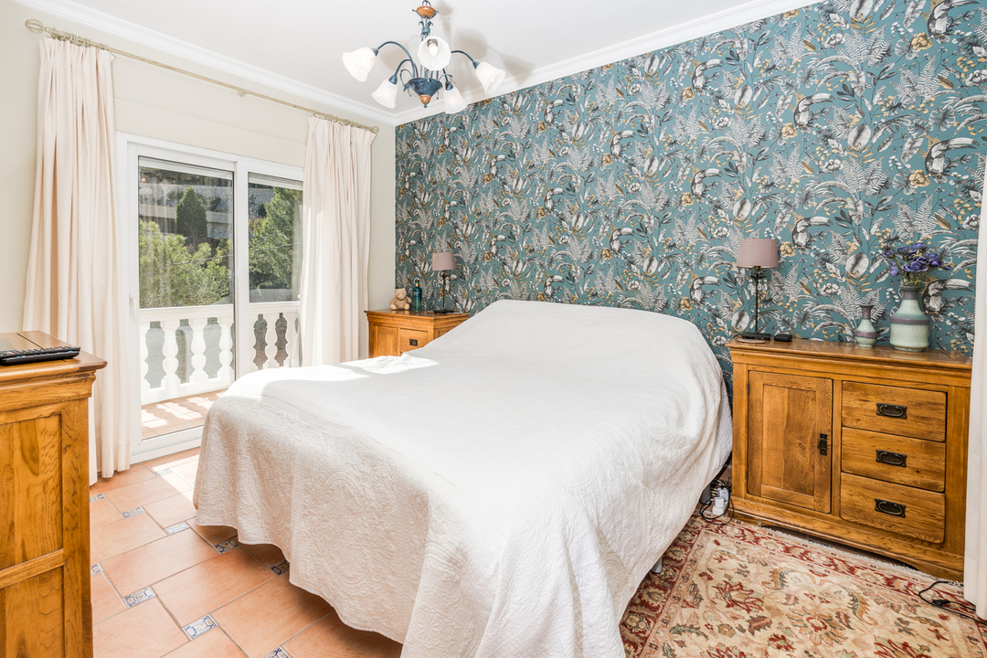 Bedroom featuring botanical wallpaper, wooden cabinetry, and terracotta tile flooring. A sliding glass door opens to a balcony with white balustrades.