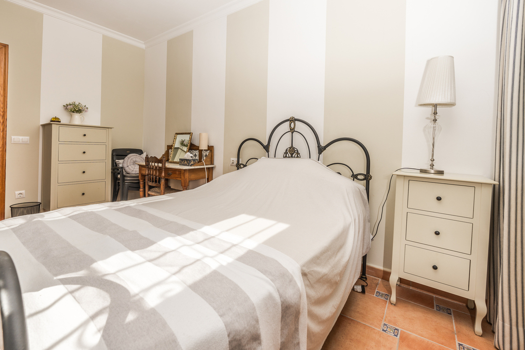Bedroom with a wrought iron headboard, striped wallpaper, and terracotta flooring. Features include a cream nightstand and a wooden vanity desk.