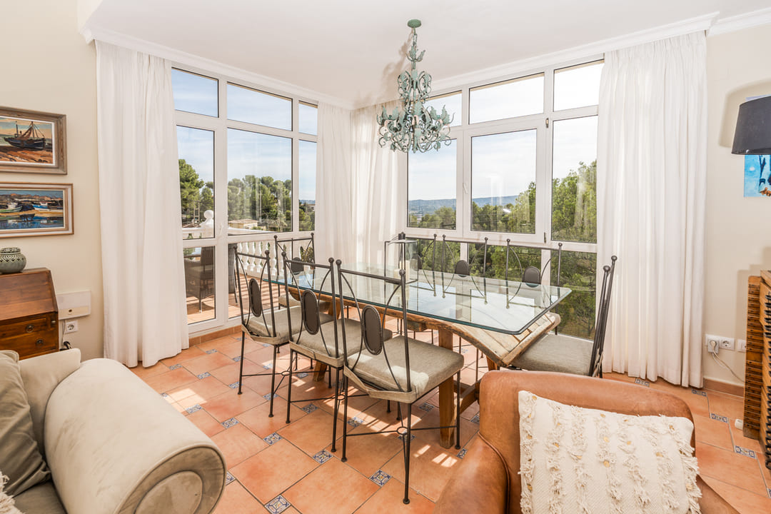 Bright dining area featuring a glass-top table, wrought iron chairs, and terracotta tiling. Large windows offer natural light and views of the surrounding greenery.