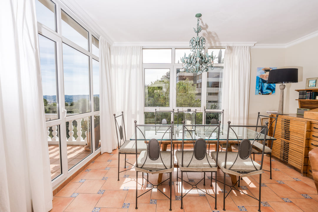 Dining area featuring a glass-top table and wrought iron chairs on terracotta tiles. Large windows provide abundant natural light and views of the surrounding landscape.