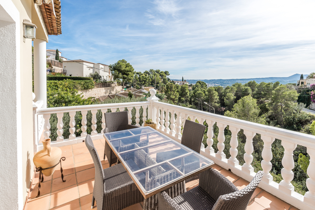 Elevated terrace featuring a glass-top dining table, wicker seating, and a classic white balustrade overlooking a pine-filled valley and distant hills.