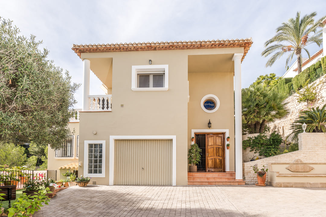 Exterior facade of a two-story villa featuring a tan finish, integrated garage, wooden entrance door, and a paved driveway surrounded by Mediterranean vegetation.