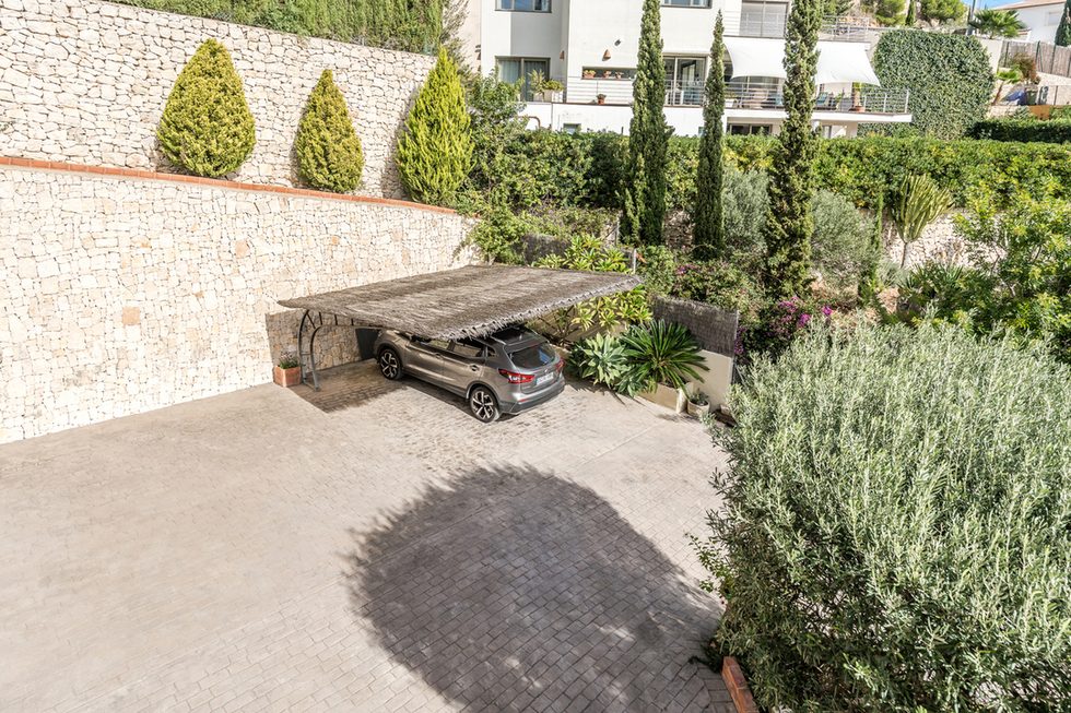 Exterior parking area featuring a paved driveway and heather-roofed carport, bordered by a dry-stone wall and manicured Mediterranean landscaping.