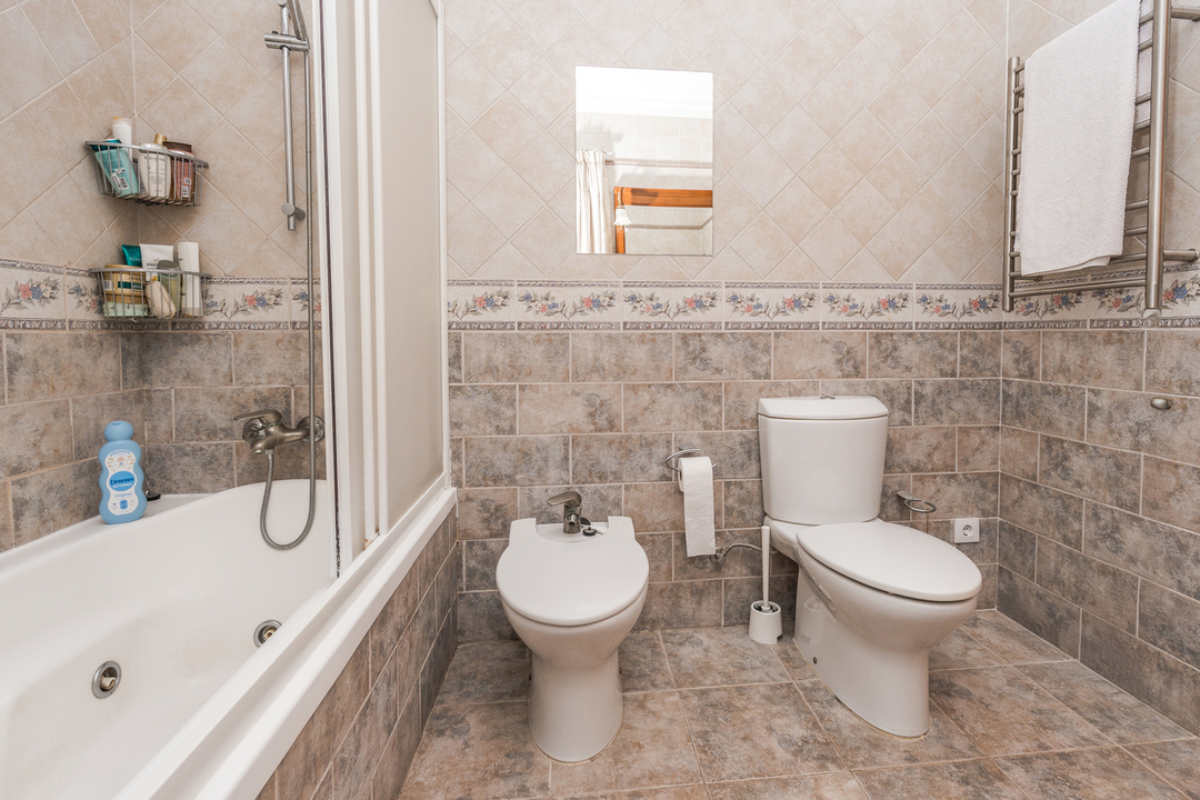 Full bathroom featuring a bathtub with glass screen, bidet, and toilet. Earth-toned wall tiles are accented by a decorative floral border and a heated towel rack.