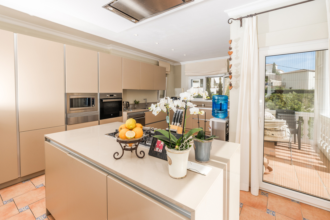 Modern kitchen featuring a beige central island with a gas hob, integrated appliances, and terracotta flooring. Large glass doors provide direct access to the terrace.