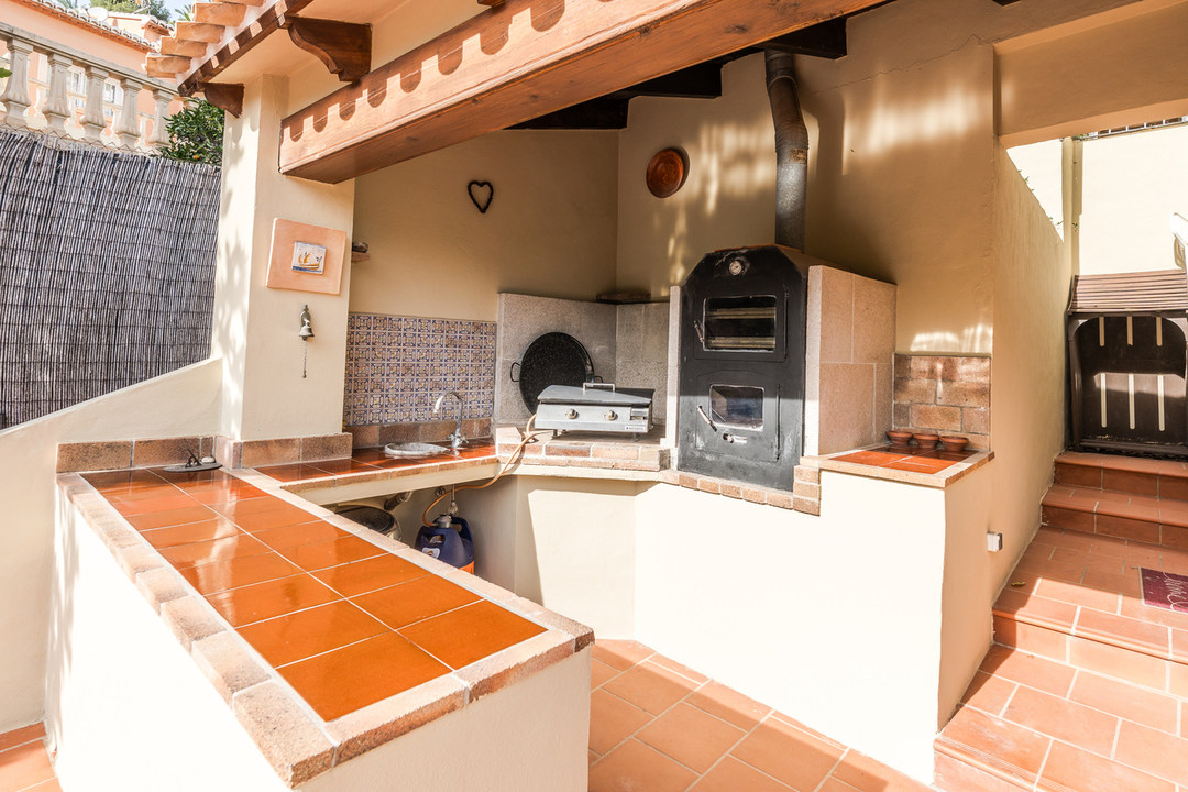 Outdoor kitchen featuring a wood-fired oven, terracotta tiled countertops, and a decorative tile backsplash under a beamed wooden porch.