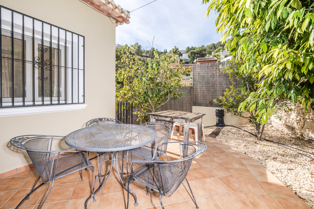 Outdoor terrace featuring terracotta tiling, a wrought-iron dining set, and a window with security bars, bordered by Mediterranean trees and gravel landscaping.
