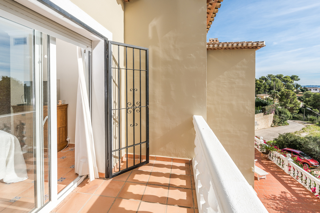 Private balcony with terracotta tiling and white balustrade, featuring a black wrought iron security gate and views of the surrounding Mediterranean landscape.