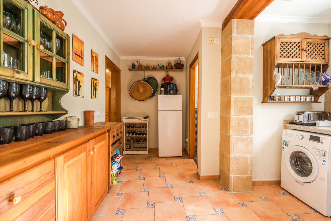 Rustic kitchen area featuring wooden cabinetry, terracotta tile flooring with blue accents, a prominent stone pillar, and integrated laundry facilities.