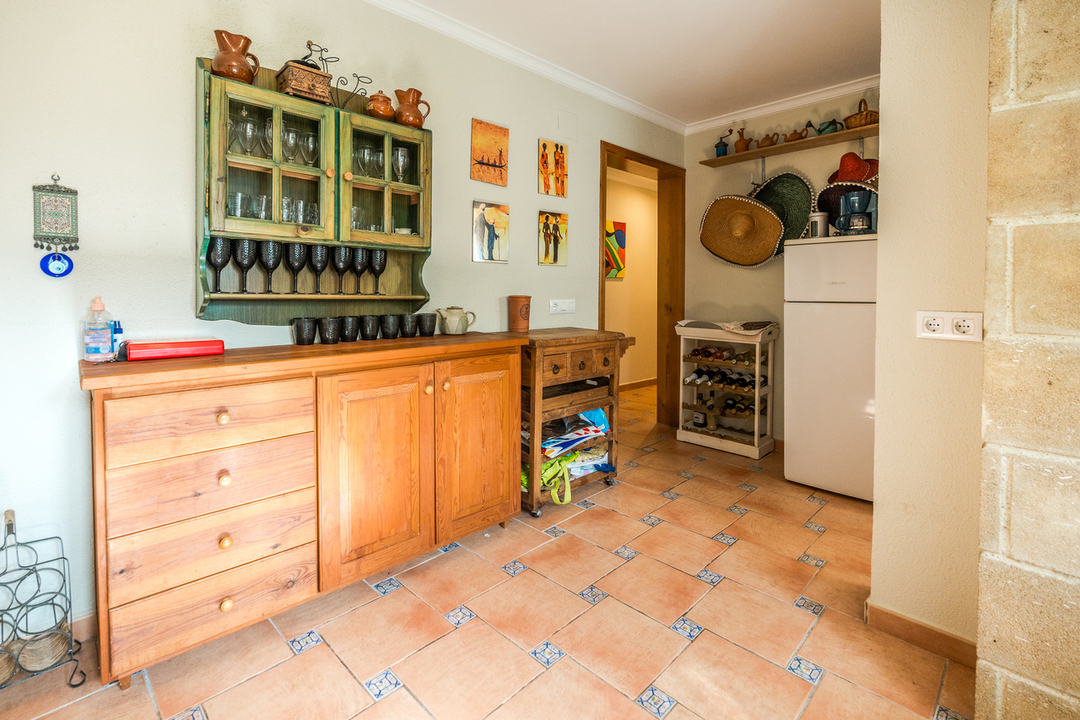 Rustic wooden sideboard and green hutch display glassware above terracotta tile flooring with decorative blue inserts. A white refrigerator and wine rack complete the space.