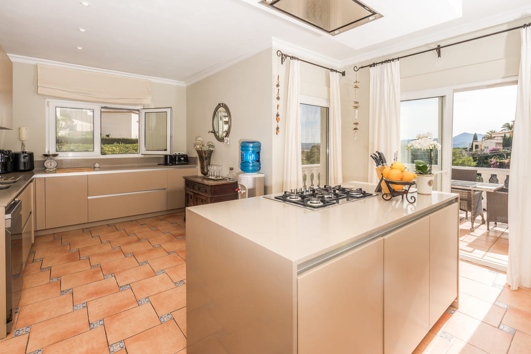 Spacious kitchen featuring a central island with a gas cooktop, beige cabinetry, and traditional terracotta floor tiles. Terrace access provides abundant natural light.