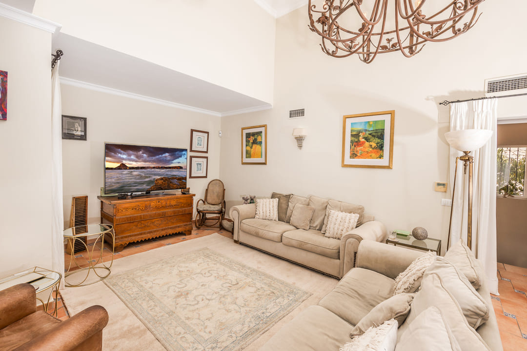 Spacious living area featuring double-height ceilings, a large wrought iron chandelier, beige sofas, and traditional terracotta tile flooring.