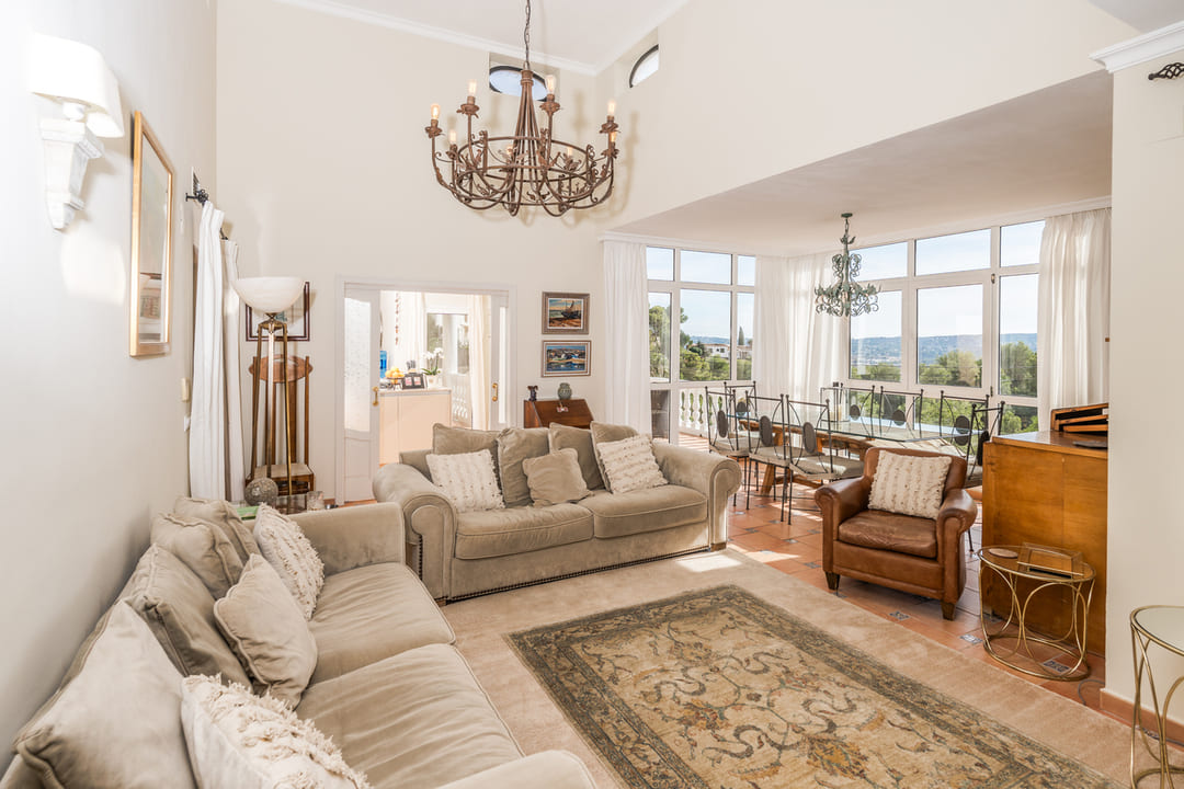 Spacious living room with high ceilings, a wrought iron chandelier, and terracotta flooring. Large windows offer natural light and views of the surrounding landscape.