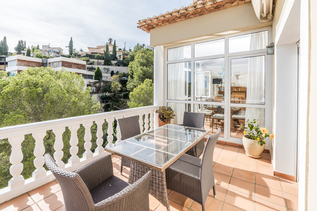 Terrace with terracotta flooring, white balustrade, and glass-top dining set. It overlooks a lush hillside with Mediterranean vegetation and neighboring villas.