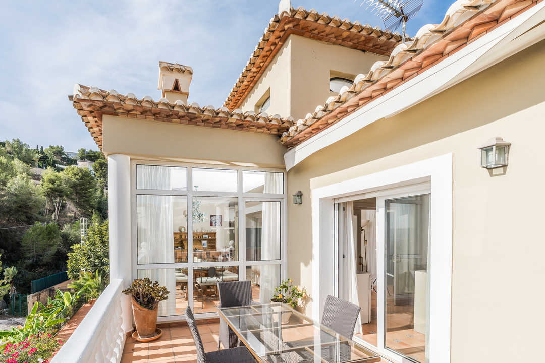 Terrace with terracotta tiling, a glass-topped dining set, and white balustrades. Large windows reveal a sunroom, set against a backdrop of Mediterranean architecture and hills.