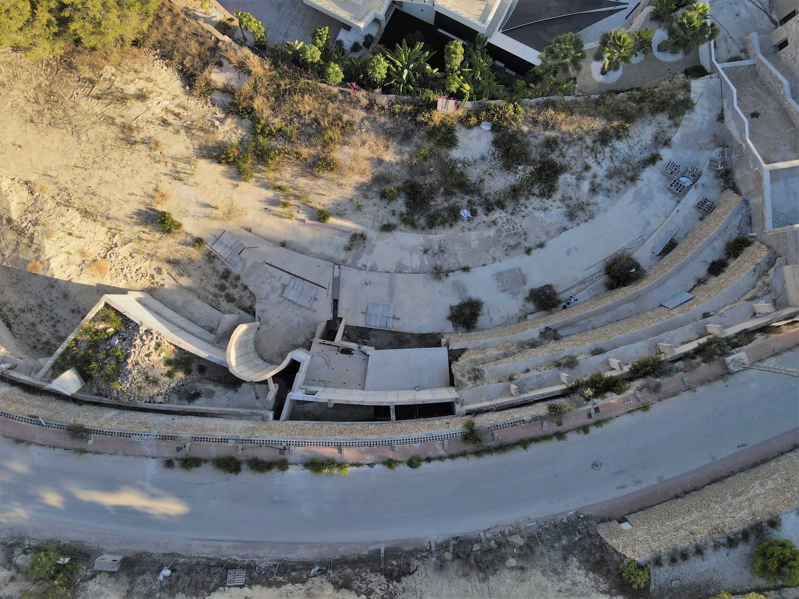 Aerial view of a terraced construction site featuring curved concrete foundations and stone retaining walls integrated into the sloped terrain.