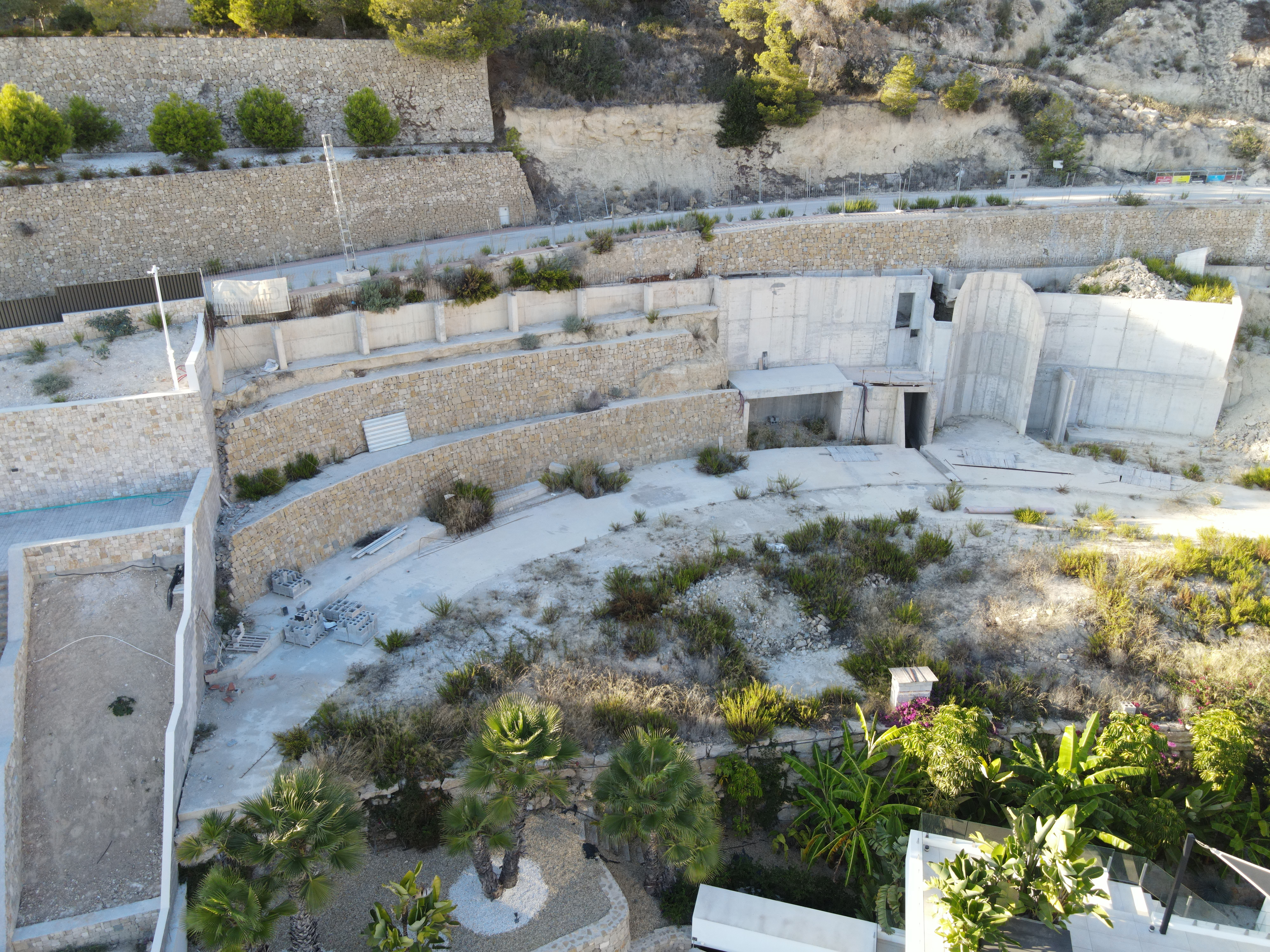 Aerial view of tiered stone retaining walls and concrete structural foundations integrated into a hillside landscape with palm trees.
