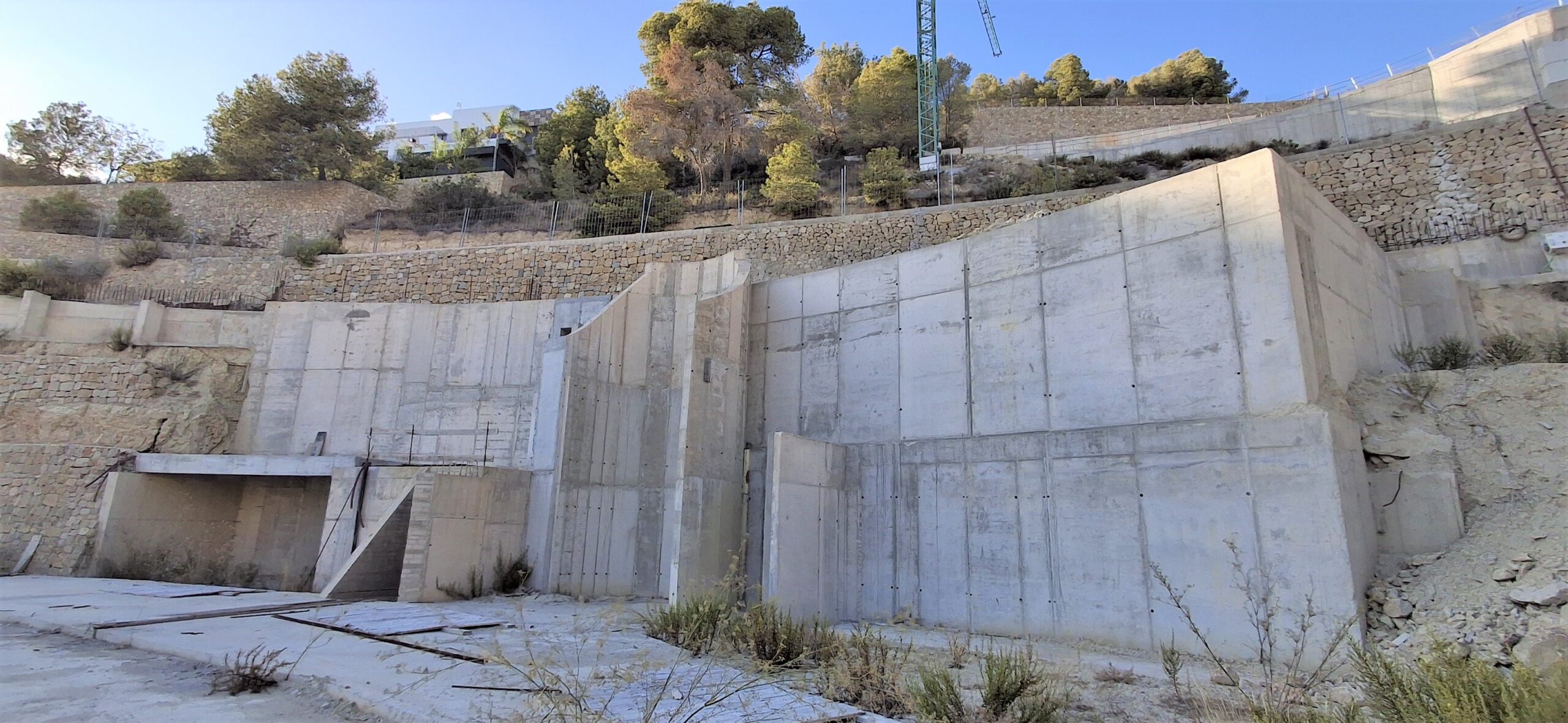 Structural view of reinforced concrete retaining walls and stone masonry terraces integrated into a steep hillside under a clear sky.